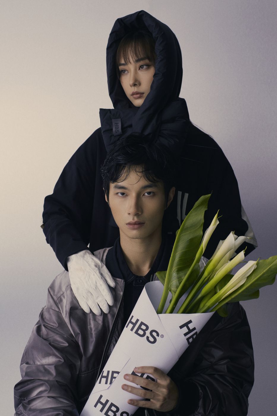 Hooded behind him, he holds the lilies